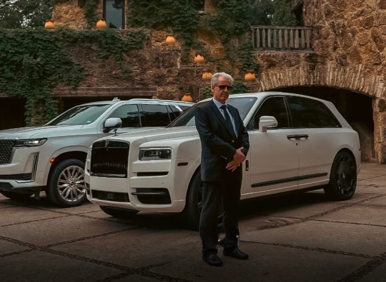 Leozen chauffeur in a dark suit standing beside a white Rolls-Royce Cullinan and a white Cadillac Escalade